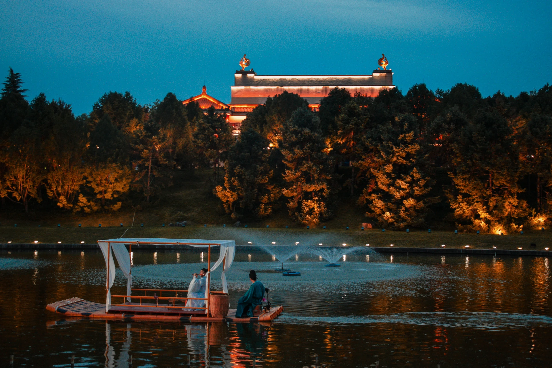 A covered boat on a lake below a brightly lit pavilion and wooded hillside