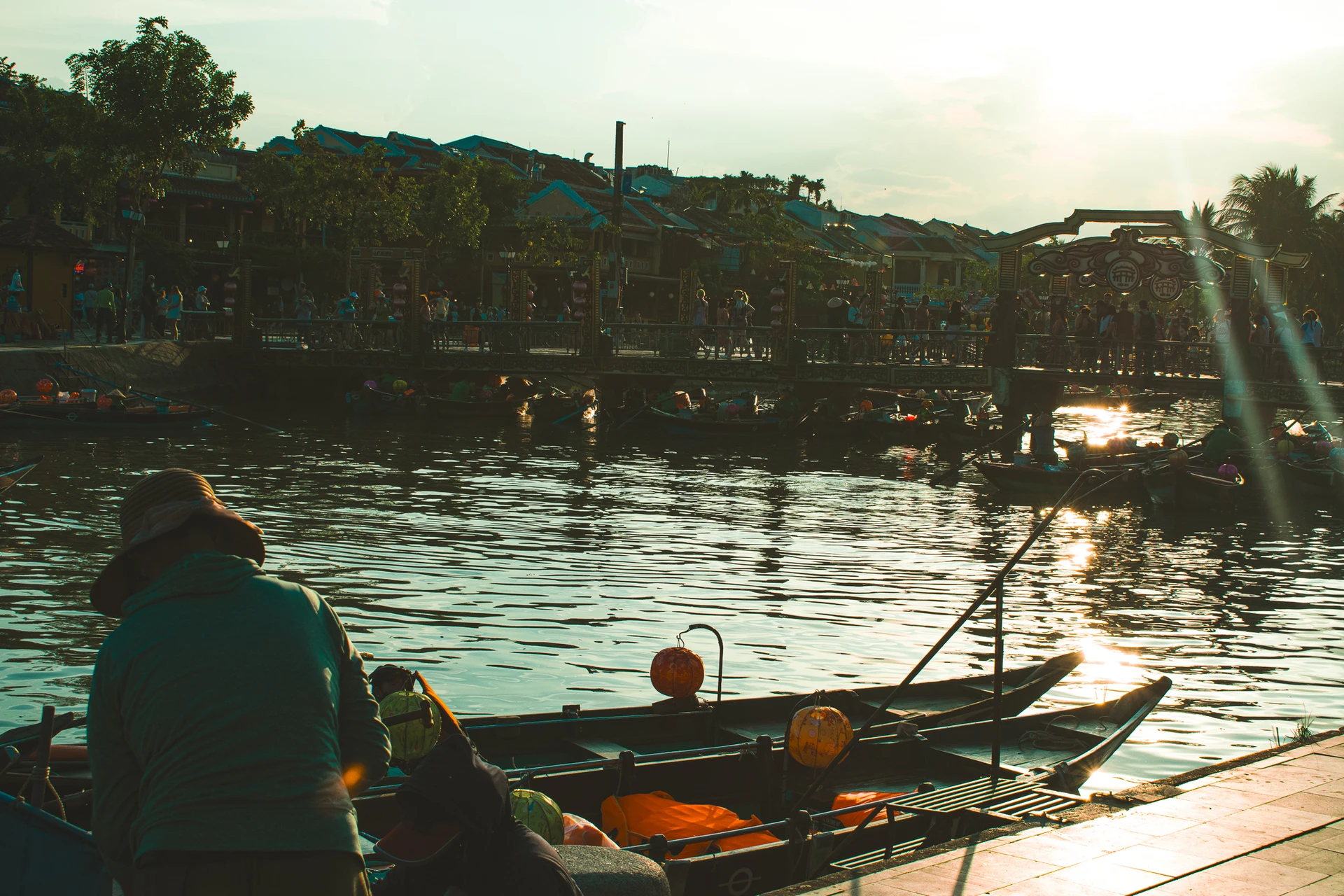 A person in a conical hat beside lantern-decorated rowing boats on a sunlit river