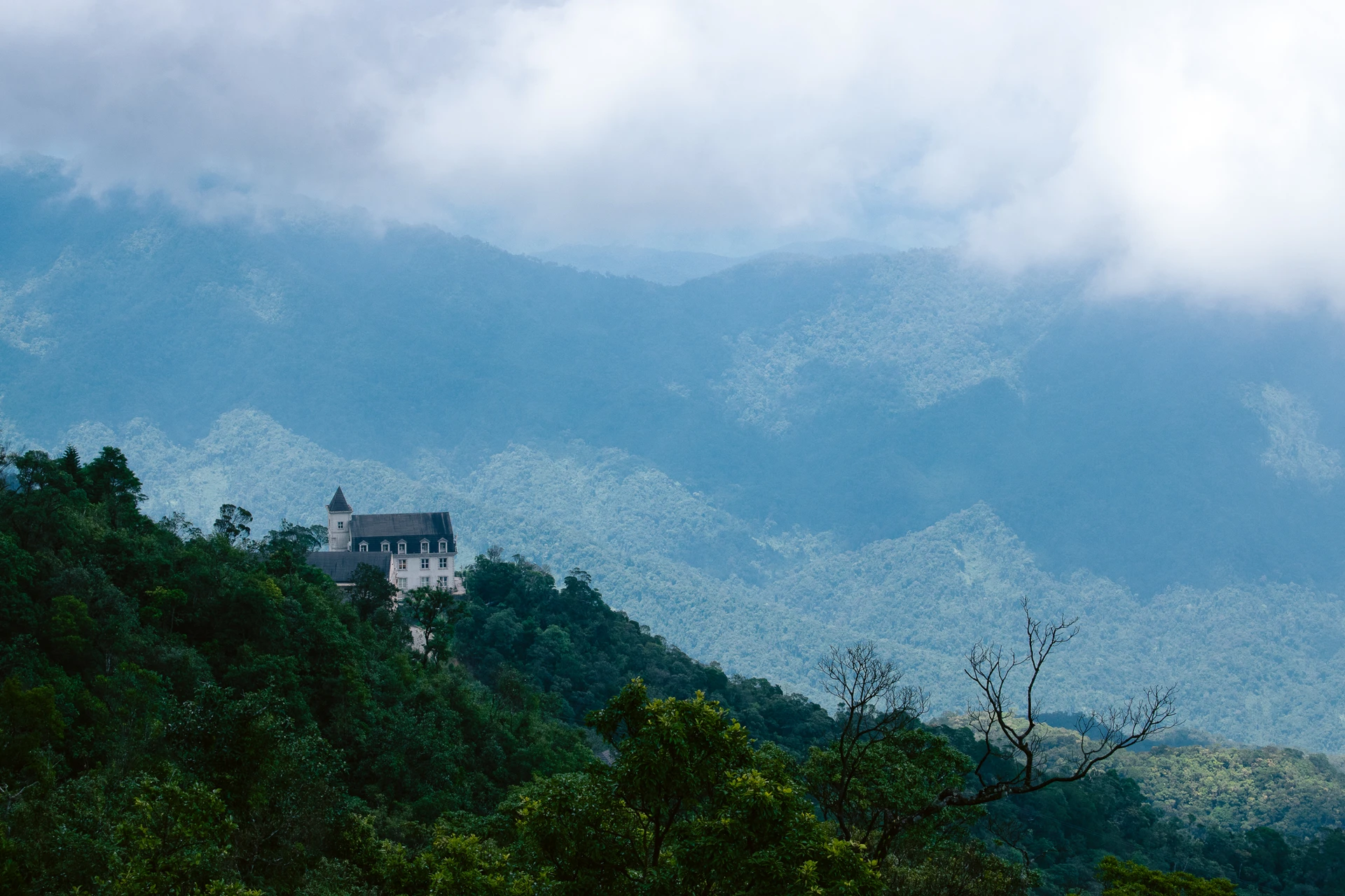 A pale building with a pointed tower on a forested hillside beneath misty mountains