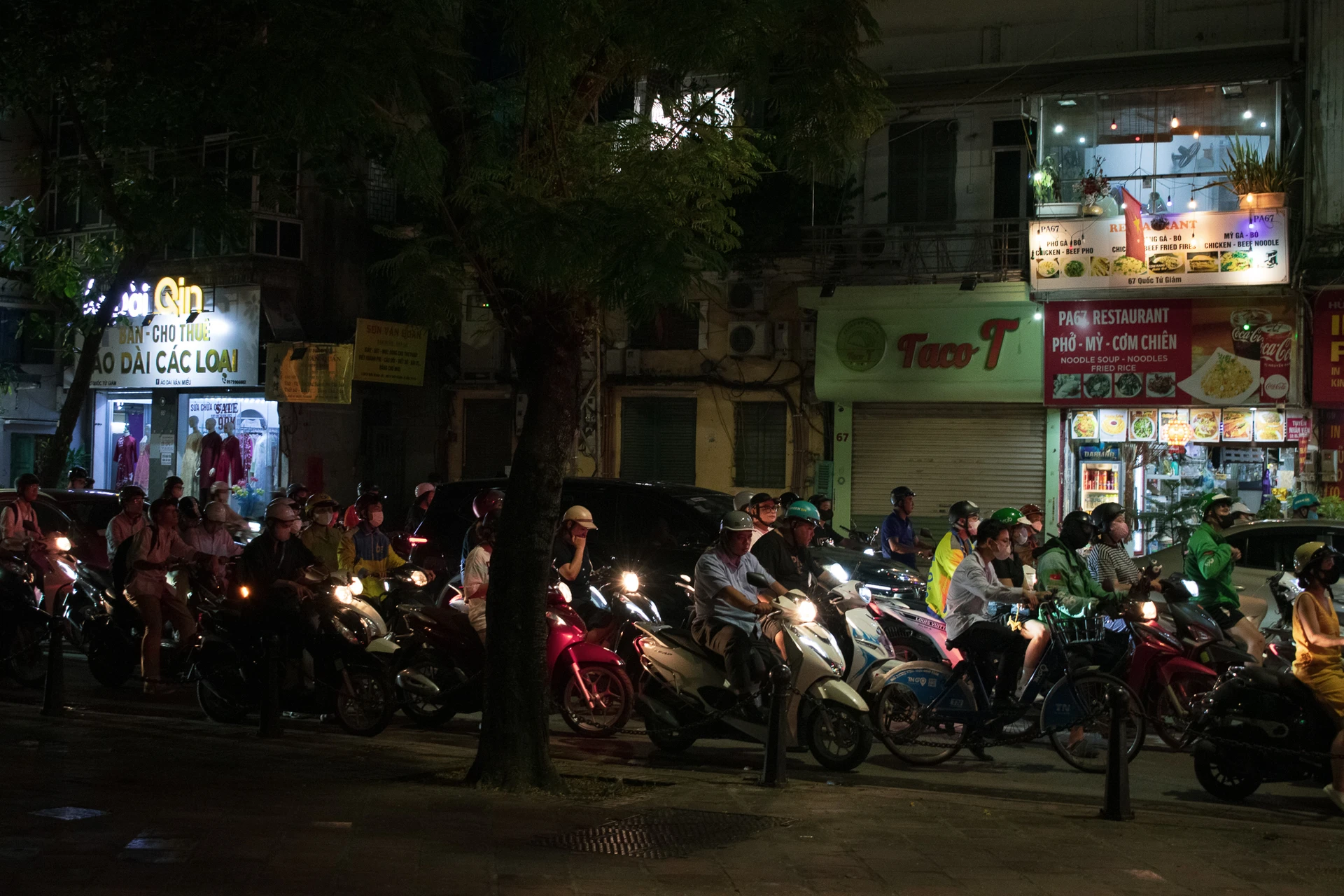 Motorbike riders gathered beneath trees and glowing shop signs on a city street at night