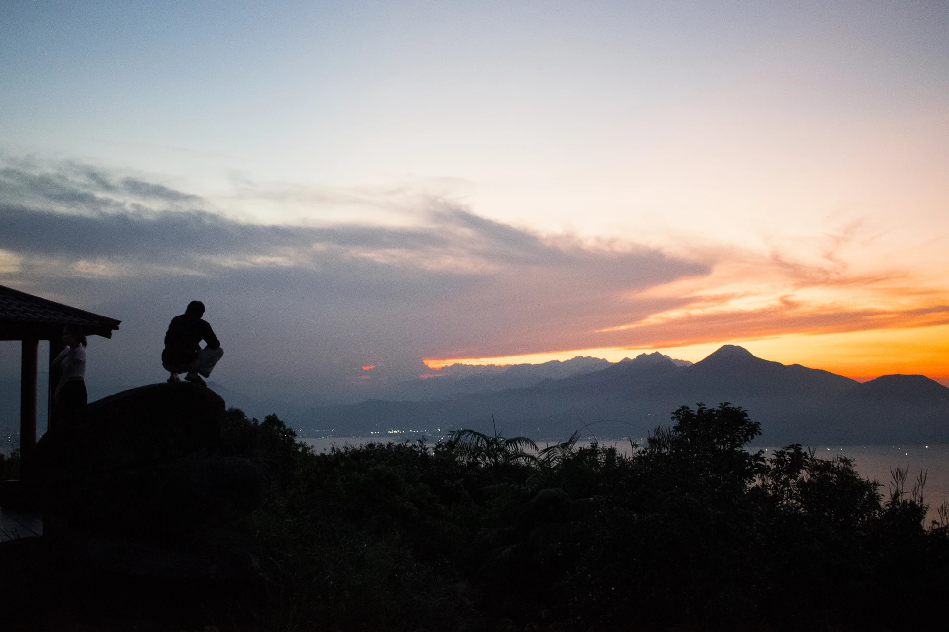 A person silhouetted on a rock overlooking water and mountain ridges beneath an orange sunset
