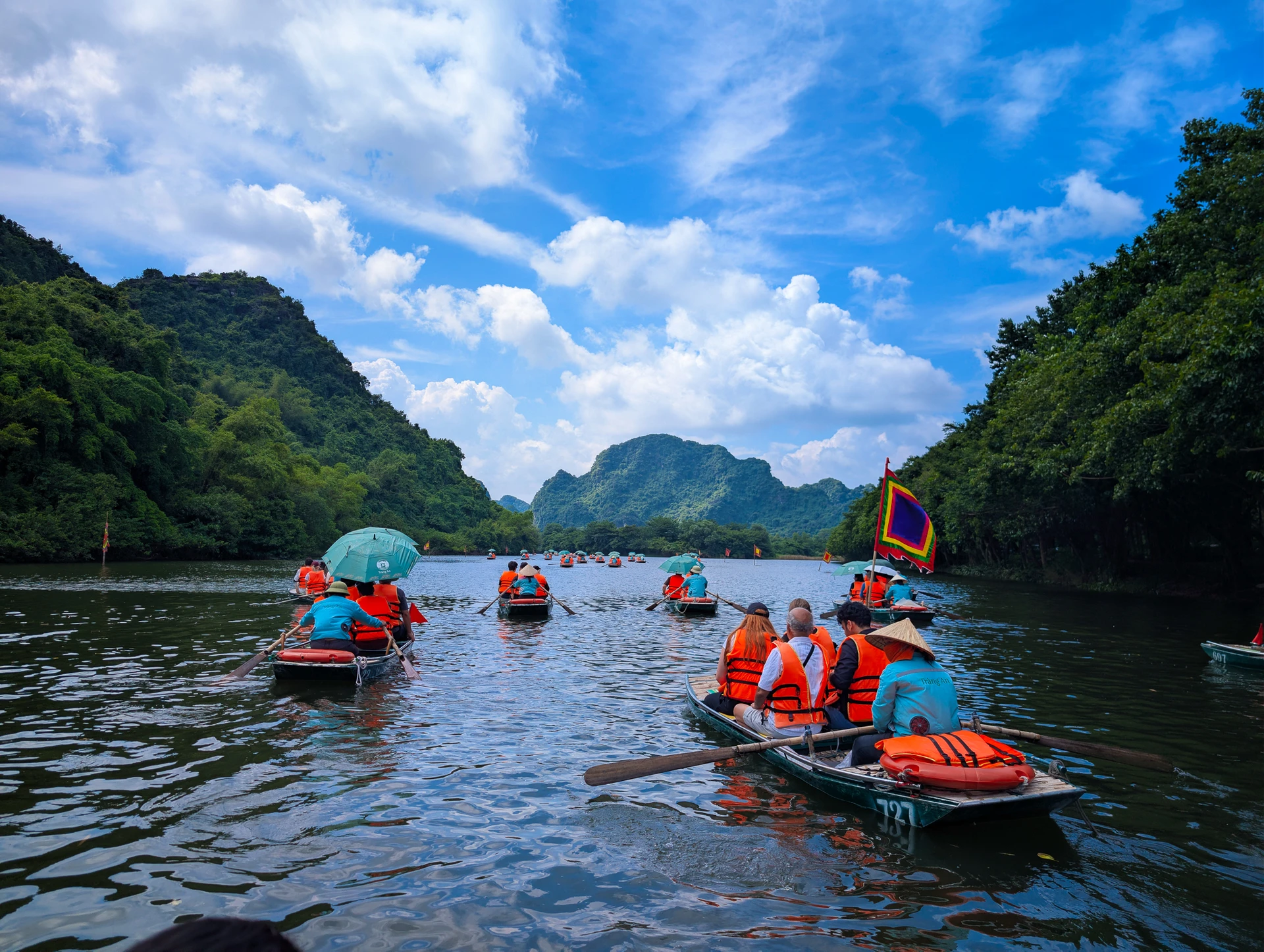 Passengers in orange life jackets travelling by rowing boat along a river between forested limestone hills