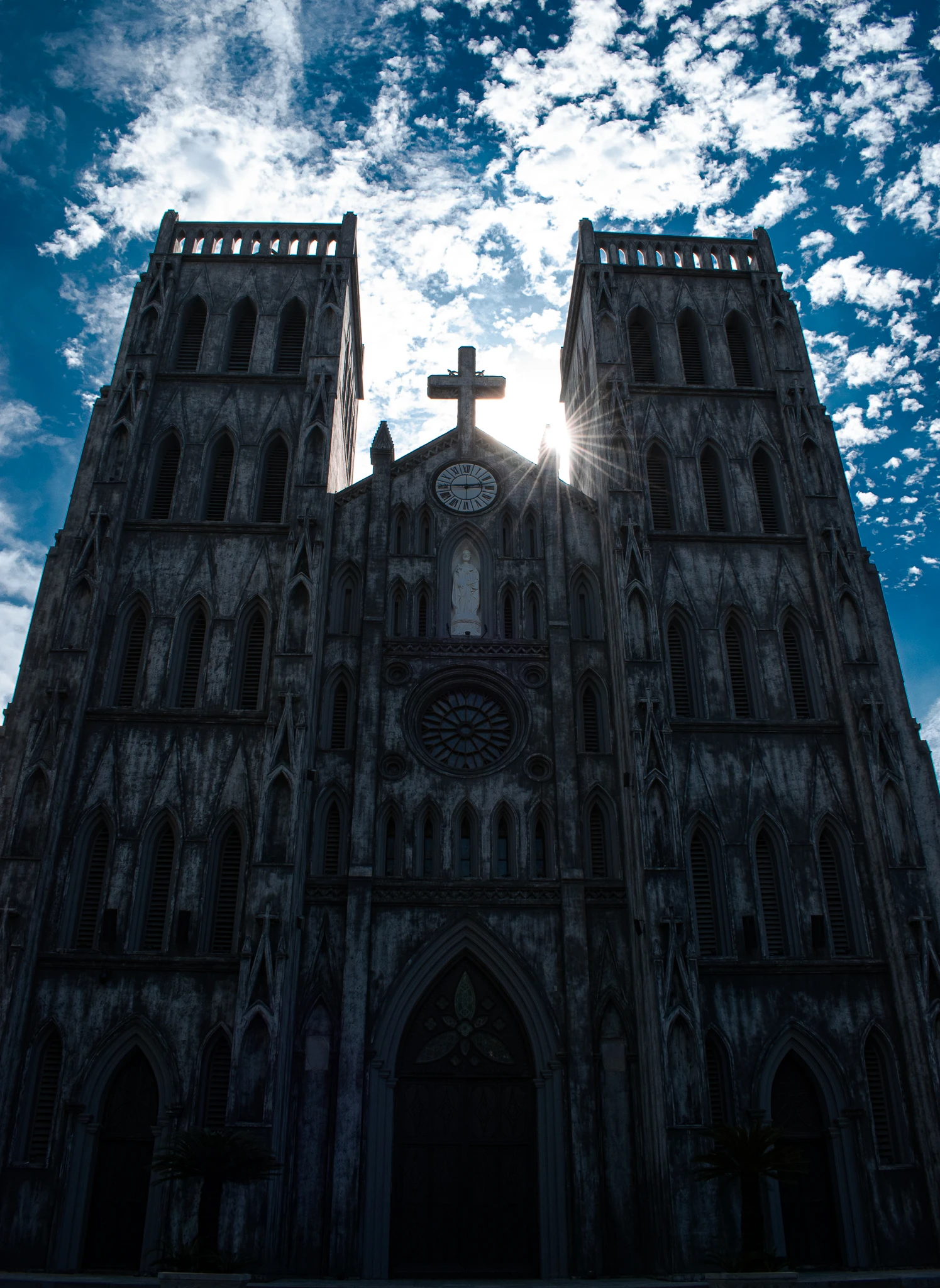Sunlight shining beside a cross between the twin towers of a dark stone cathedral
