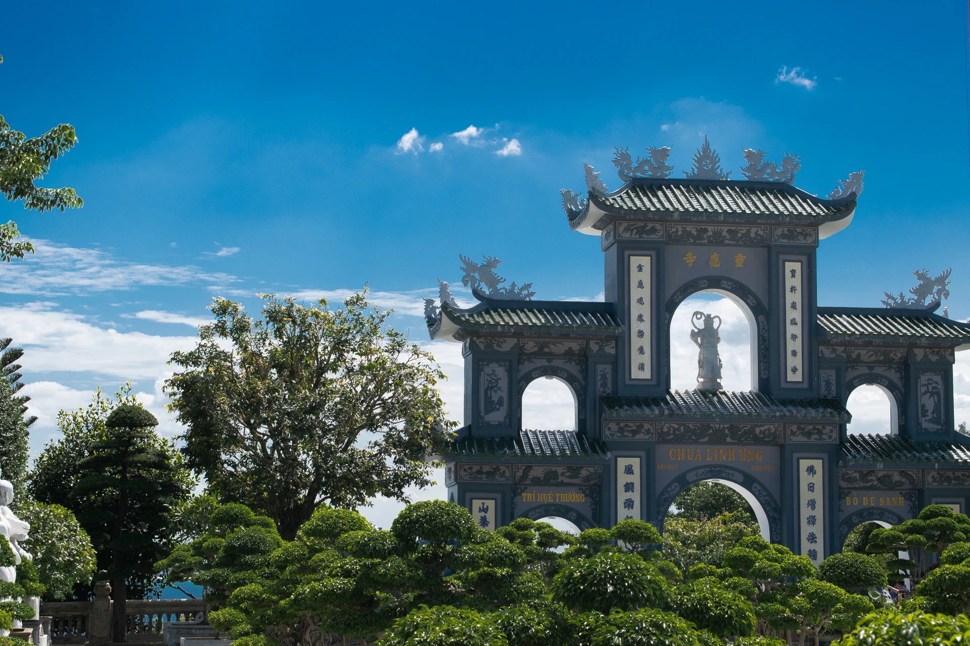 An ornate temple gateway with curved tiled roofs and a white statue above neatly clipped trees